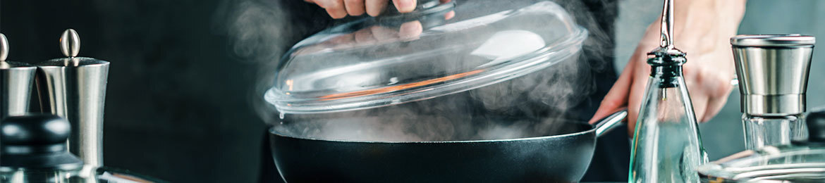 Ein Koch hebt den Glasdeckel einer Pfanne an, Dampf strömt aus.